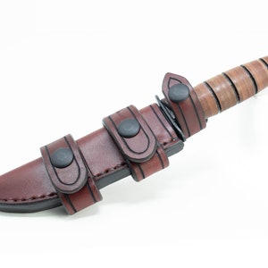 May include: A brown leather knife sheath with black stitching and snaps. The sheath has three straps for securing the knife. The knife is not visible in the image.