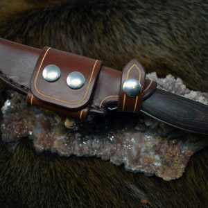 May include: A brown leather knife sheath with a tan stitching detail and silver snaps. The sheath is holding a small knife with a dark wood handle.