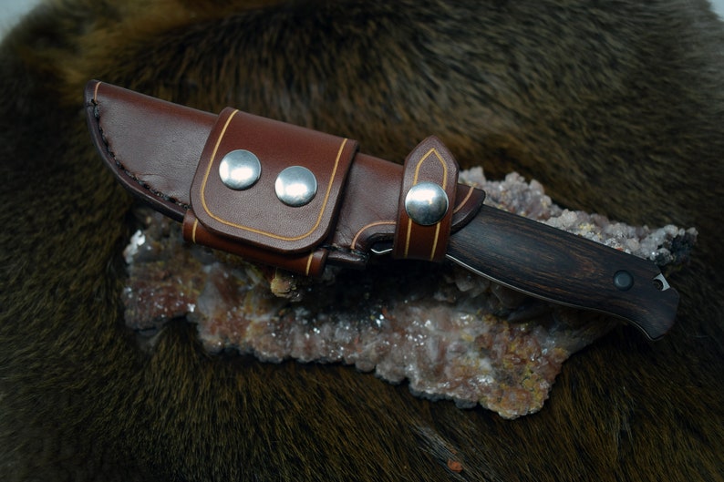 May include: A brown leather knife sheath with a tan stitching detail and silver snaps. The sheath is holding a small knife with a dark wood handle.