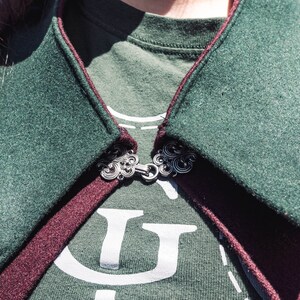 Puede incluir: Primer plano de una capa con capucha verde y burdeos con un broche de plata. La capa est&aacute; hecha de una tela suave y texturizada.