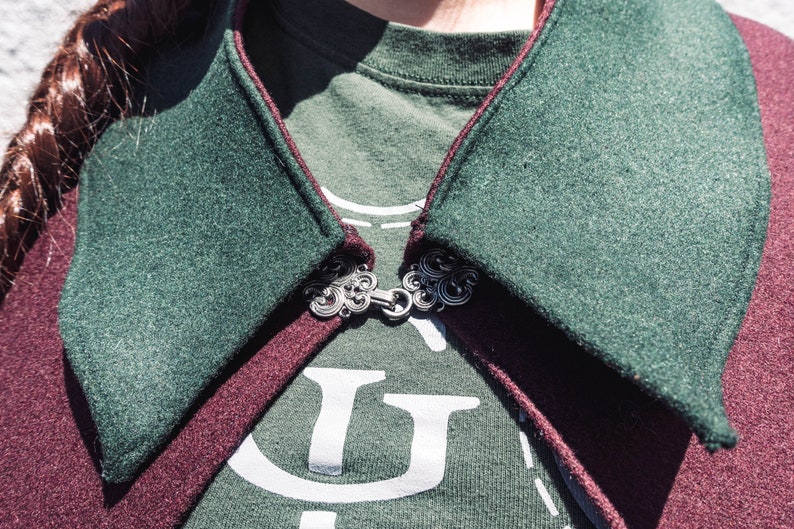 Puede incluir: Primer plano de una capa con capucha verde y burdeos con un broche de plata. La capa est&aacute; hecha de una tela suave y texturizada.