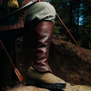 May include: A pair of dark brown leather gaiters worn over light olive green shoes. The gaiters feature a smooth, textured surface and are secured with side lacing. The footwear has a thick, black lugged rubber sole, providing a sturdy base.