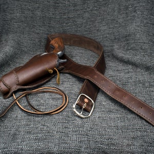 Może przedstawiać: Brązowy sk&oacute;rzany holster i pasek ze srebrną klamrą. Holster jest zaprojektowany do trzymania pistoletu na biodrze.