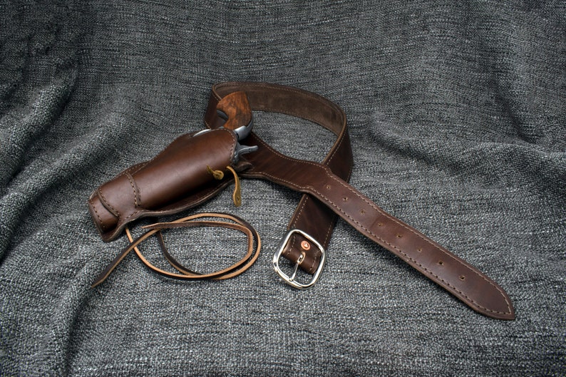 Może przedstawiać: Brązowy sk&oacute;rzany holster i pasek ze srebrną klamrą. Holster jest zaprojektowany do trzymania pistoletu na biodrze.