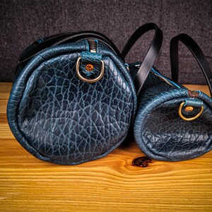 Puede incluir: Dos bolsas de viaje de cuero azul con asas negras y herrajes dorados. La bolsa m&aacute;s grande est&aacute; en primer plano y la m&aacute;s peque&ntilde;a est&aacute; detr&aacute;s.