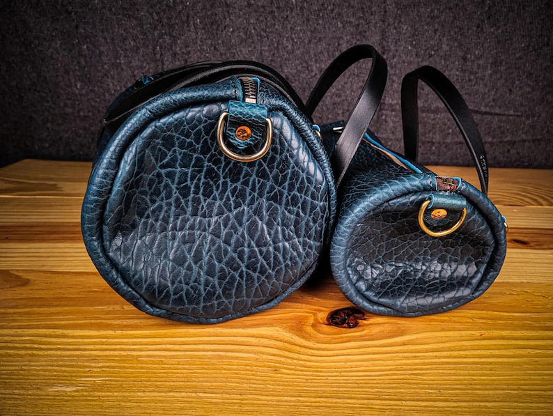 Puede incluir: Dos bolsas de viaje de cuero azul con asas negras y herrajes dorados. La bolsa m&aacute;s grande est&aacute; en primer plano y la m&aacute;s peque&ntilde;a est&aacute; detr&aacute;s.