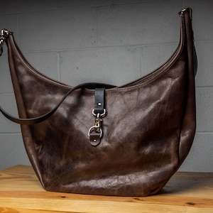 Puede incluir: Un bolso de cuero marrón con una correa larga y un cierre de metal. El bolso tiene una forma curva y un aspecto desgastado.