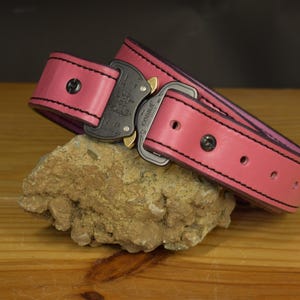 K&ouml;nnte beinhalten: Ein pinkfarbener Lederg&uuml;rtel mit schwarzen N&auml;hten an den Kanten und einer schwarzen Metallschnalle im Cobra-Stil. Der G&uuml;rtel verf&uuml;gt &uuml;ber eine Reihe von Einstelll&ouml;chern und dunkle Metallnieten, was eine robuste Konstruktion mit einem leuchtenden Finish zeigt.