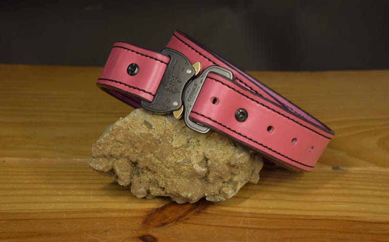 K&ouml;nnte beinhalten: Ein pinkfarbener Lederg&uuml;rtel mit schwarzen N&auml;hten an den Kanten und einer schwarzen Metallschnalle im Cobra-Stil. Der G&uuml;rtel verf&uuml;gt &uuml;ber eine Reihe von Einstelll&ouml;chern und dunkle Metallnieten, was eine robuste Konstruktion mit einem leuchtenden Finish zeigt.