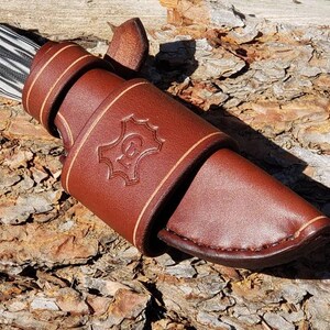 May include: A brown leather knife sheath with a decorative stitching detail and a stamped logo. The sheath is designed to hold a knife with a black and white patterned handle.