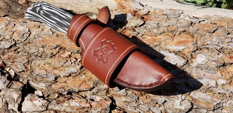 May include: A brown leather knife sheath with a decorative stitching detail and a stamped logo. The sheath is designed to hold a knife with a black and white patterned handle.
