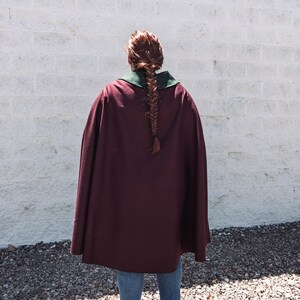 Puede incluir: Una persona lleva una capa granate con cuello verde. La capa es larga y fluida, y la persona est&aacute; de pie frente a una pared de ladrillo blanco.