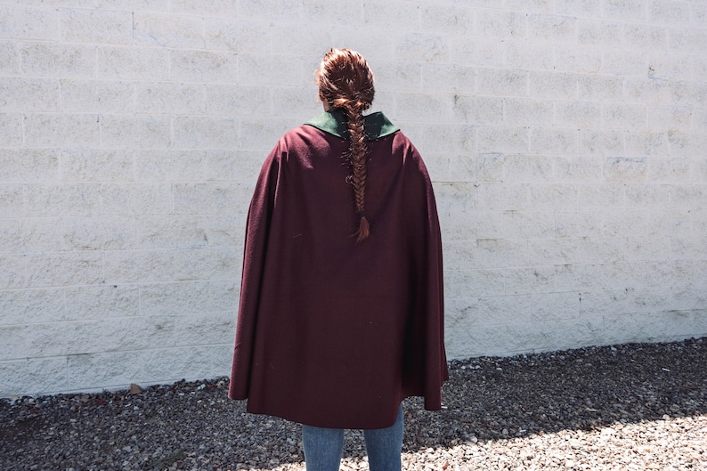 Puede incluir: Una persona lleva una capa granate con cuello verde. La capa es larga y fluida, y la persona est&aacute; de pie frente a una pared de ladrillo blanco.