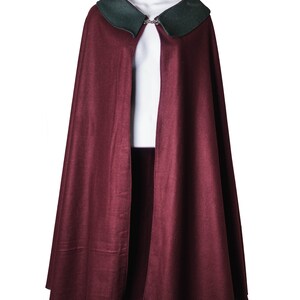 Puede incluir: Una capa de lana color burdeos con cuello de terciopelo verde y cierre de plata.