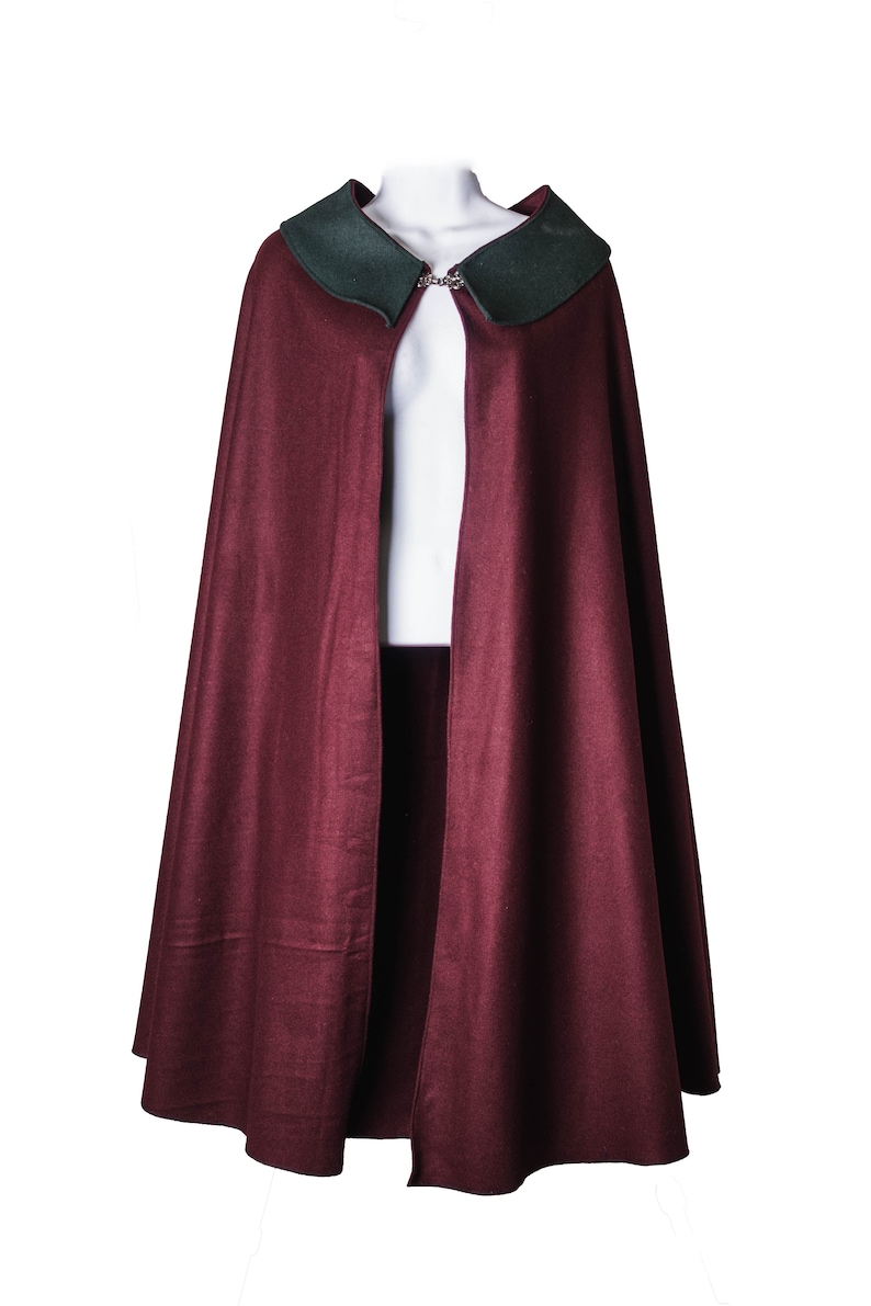 Puede incluir: Una capa de lana color burdeos con cuello de terciopelo verde y cierre de plata.