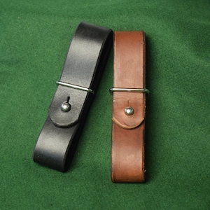 Puede incluir: Dos correas de cuero con herrajes metálicos. Una correa es negra, la otra marrón. Cada correa tiene un anillo metálico plateado y un cierre de botón plateado. Las correas se muestran sobre un fondo verde.