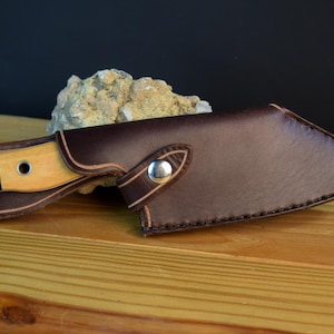 May include: A brown leather knife sheath with a snap closure and a stitched edge. The sheath is designed to fit a knife with a wooden handle.