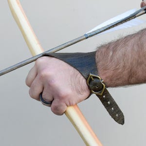 以下が含まれることがあります： 真鍮のバックルで留められた、濃い茶色の革製アーチェリーアームガード。アームガードは前腕に着用し、弓弦から保護します。矢が木製の弓に番えられています。