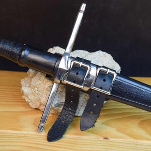 May include: A medieval sword with a silver blade and a black leather-wrapped handle. The sword is sheathed in a black leather scabbard, secured by two leather straps with silver buckles. The sword rests on a light-colored stone.