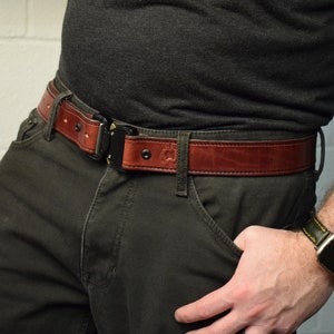 K&ouml;nnte beinhalten: Ein brauner Lederg&uuml;rtel mit einer schwarzen Metallschnalle mit Schnellverschluss. Der G&uuml;rtel ist mit sichtbaren N&auml;hten an den Kanten versehen und enth&auml;lt ein kleines gepr&auml;gtes Logo auf dem Lederriemen in der N&auml;he des Schnallenmechanismus.