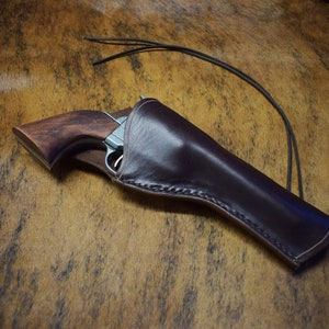 Può includere: Un fodero in pelle marrone con un bordo cucito, che contiene una pistola d'argento con un'impugnatura in legno. Il fodero ha una cinghia in pelle.