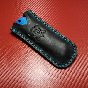 Peut inclure: Un étui à couteau en cuir noir avec des détails de couture bleus et un logo en cuir. L'étui est conçu pour contenir un petit couteau de poche.