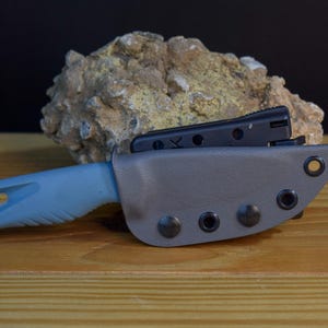 Puede incluir: Un cuchillo azul y gris con una funda negra. El cuchillo está en posición horizontal con la hoja orientada hacia el lado derecho de la imagen. La funda tiene un cierre de presión y está hecha de un material texturizado. El cuchillo está sobre una superficie de madera.