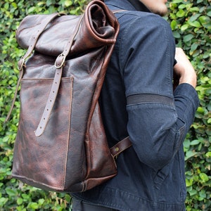 May include: A brown leather backpack with a roll-top closure and adjustable straps. The backpack has a large pocket on the front and is made of distressed leather.