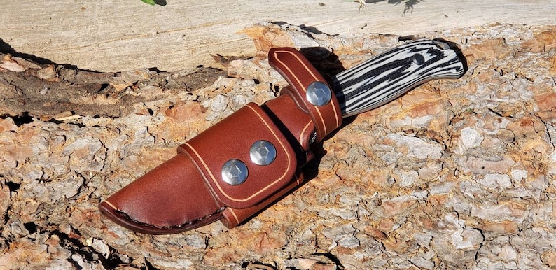 May include: A brown leather knife sheath with a snap closure and three metal rivets. The sheath is designed to hold a knife with a black and white patterned handle.