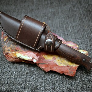 May include: A brown leather sheath with a light brown stitching detail holds a dark wood handled knife with a silver rivet. The knife is resting on a red, yellow, and white rock.