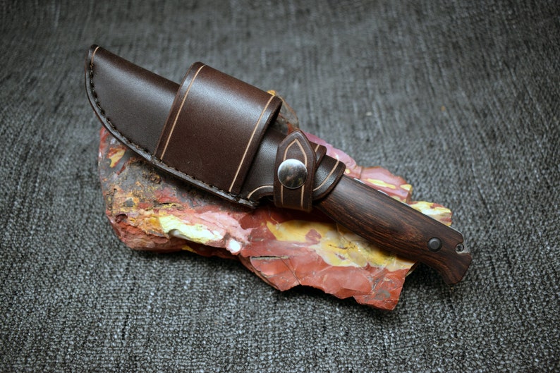 May include: A brown leather sheath with a light brown stitching detail holds a dark wood handled knife with a silver rivet. The knife is resting on a red, yellow, and white rock.