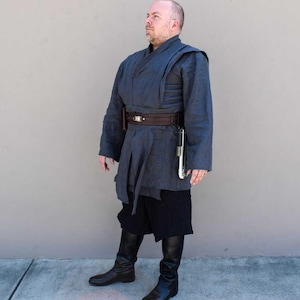 May include: A dark blue costume with a wrap-style top, a brown belt, and black shorts. The costume includes a silver and white prop and black boots. The outfit is reminiscent of a Jedi or similar character.