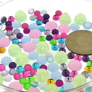 May include: A clear glass bowl filled with colorful, assorted-sized, flat-backed rhinestones. The rhinestones are in shades of pink, green, blue, purple, and silver. A U.S. quarter is in the center of the bowl for size reference.