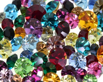 May include: A large assortment of colorful, faceted glass rhinestones, some with a metallic finish, are scattered on a white surface. A US quarter is included for scale.