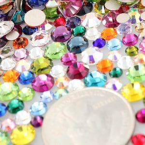 May include: A collection of colorful rhinestones in various sizes and shapes, including round and oval. The rhinestones are in shades of pink, purple, green, blue, orange, and yellow. A US quarter is included for size reference.