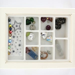 May include: A cream-colored organizer filled with various craft embellishments. The assortment includes colorful rhinestones, star-shaped sequins, heart-shaped gems, and other decorative elements. Ideal for jewelry making and craft projects.