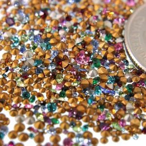 May include: A close-up of a large quantity of small, colorful, faceted rhinestones with gold-colored bases. The rhinestones are scattered on a white surface, and a US quarter is included for scale.
