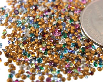 May include: A close-up of a large quantity of small, colorful, faceted rhinestones with gold-colored bases. The rhinestones are scattered on a white surface, and a US quarter is included for scale.