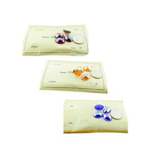 May include: Three pieces of paper with iridescent glass cabochons in various colors. The paper has the words "Bombe" and "unfoiled" printed on it. The cabochons are labeled "ruby", "topaz", and "sapphire".