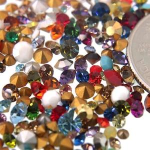 May include: A variety of colorful rhinestones and gold-toned metal embellishments scattered on a white surface. The rhinestones are in various shapes and sizes, including round, square, and oval. A US quarter is included for scale.