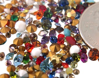 May include: A variety of colorful rhinestones and gold-toned metal embellishments scattered on a white surface. The rhinestones are in various shapes and sizes, including round, square, and oval. A US quarter is included for scale.