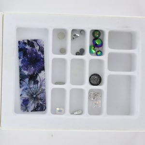 May include: A white divided tray filled with various craft supplies. Includes a floral patterned strip in shades of blue and purple, along with small iridescent gems, beads, and charms. The tray is designed for organizing small items.
