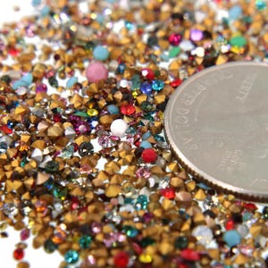May include: A close-up of a variety of colorful rhinestones and gold-toned metal studs. The rhinestones are in various shapes and sizes, including round, square, and star-shaped. A US quarter is included for scale.