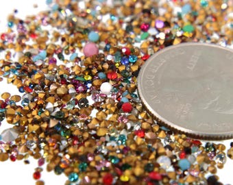 May include: A close-up of a variety of colorful rhinestones and gold-toned metal studs. The rhinestones are in various shapes and sizes, including round, square, and star-shaped. A US quarter is included for scale.