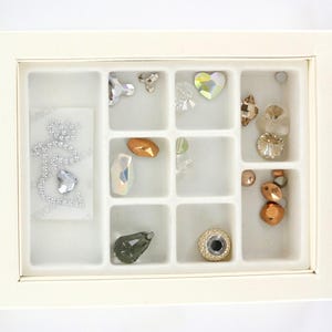 May include: A cream-colored organizer box with a grid of compartments, each holding various sparkling rhinestones and embellishments. The rhinestones are in various shapes, including hearts, ovals, and round, and in colors like clear, gold, and iridescent.