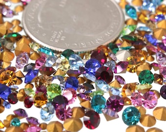 May include: A variety of colorful rhinestones and gold-toned nail studs. The rhinestones are in various shapes and sizes, including round, square, and oval. The gold-toned nail studs are small and have a pointed tip.
