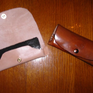 May include: Two leather cases, one open and one closed, are displayed on a wooden surface. The open case is light pink with a black interior and a gold snap closure. The closed case is brown with a black snap closure.