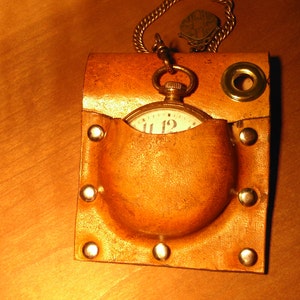 Puede incluir: Un portador de reloj de bolsillo de cuero marrón con una cadena dorada y un reloj de bolsillo con una esfera blanca y números negros.