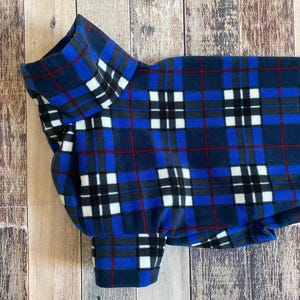 May include: A blue plaid dog sweater with a high collar and sleeves. The pattern includes navy, red, white, and gray squares and lines. The sweater is made of a soft fleece material.