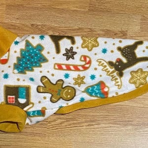 Op de afbeelding: Een hondenjas met een feestelijk kerstpatroon. De jas heeft een witte achtergrond met turquoise kerstbomen, peperkoekenmannen en rendieren. De rand en manchetten zijn mosterdgeel.